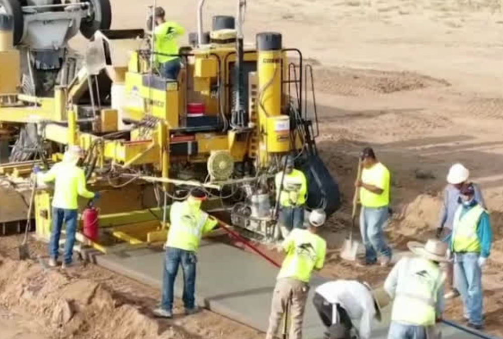 Construction crew operating a curb and gutter machine while workers finish freshly poured concrete along a roadway construction site.