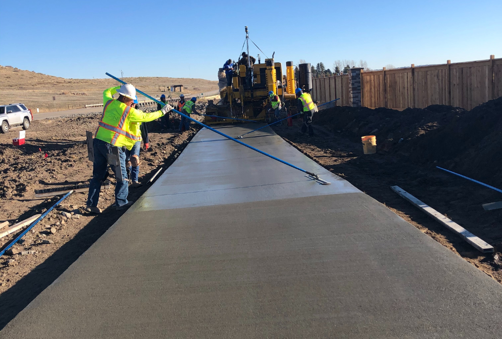 Construction crew smoothing and finishing freshly poured concrete pavement using long-handled screeds while a paving machine operates ahead.