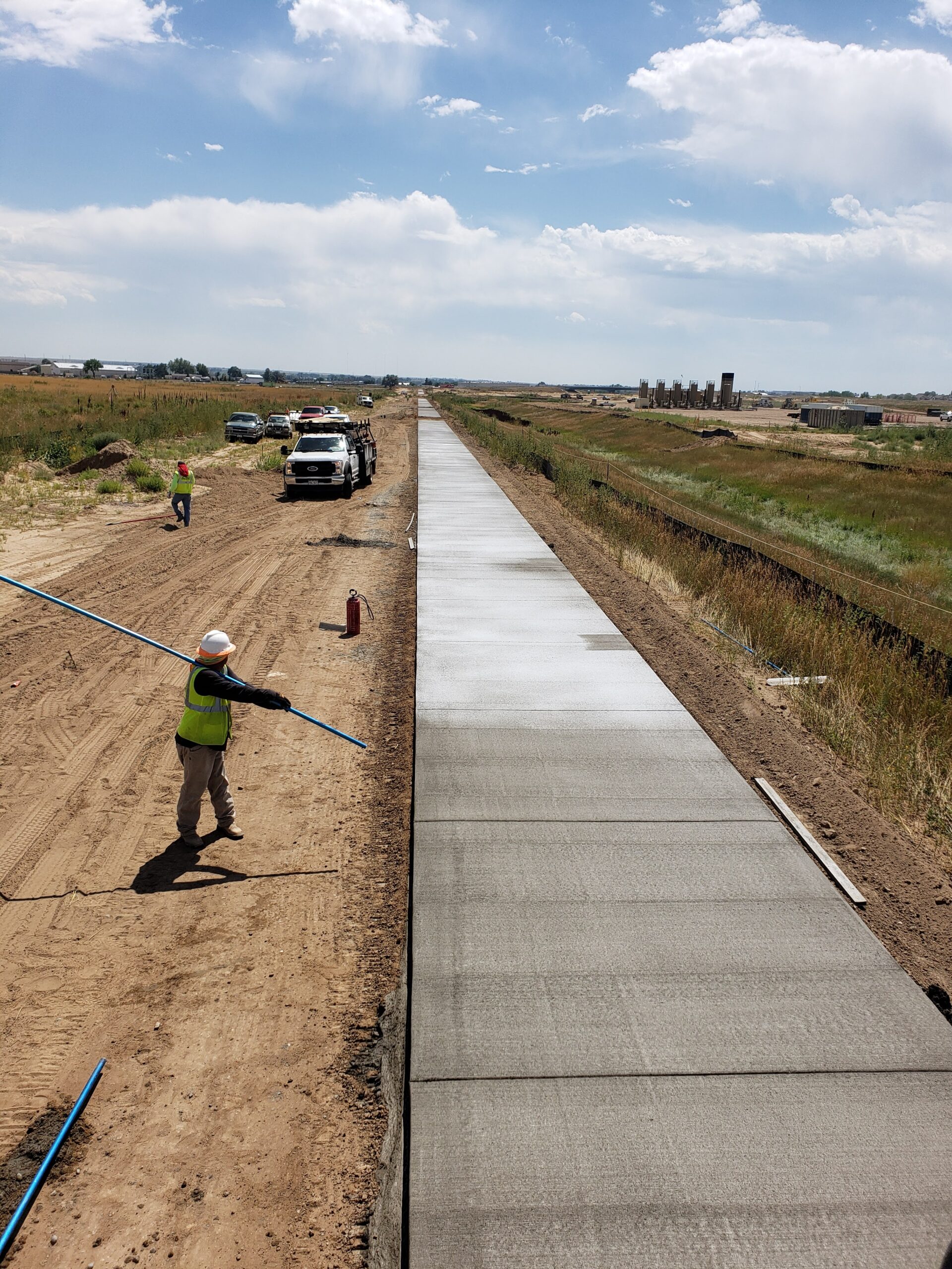Construction worker finishing a long stretch of newly poured concrete sidewalk while work trucks and crew members line a dirt access road beside the project.