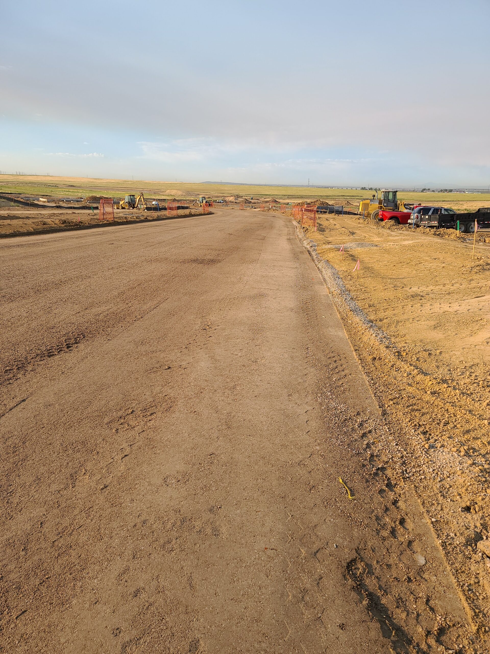 Graded dirt roadway under construction with construction fencing, equipment, and trucks along a rural development site.