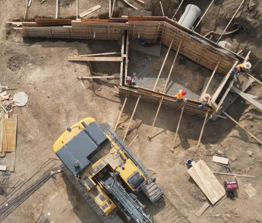 Aerial view of construction workers assembling wooden formwork for a concrete structure beside heavy equipment at an active job site.
