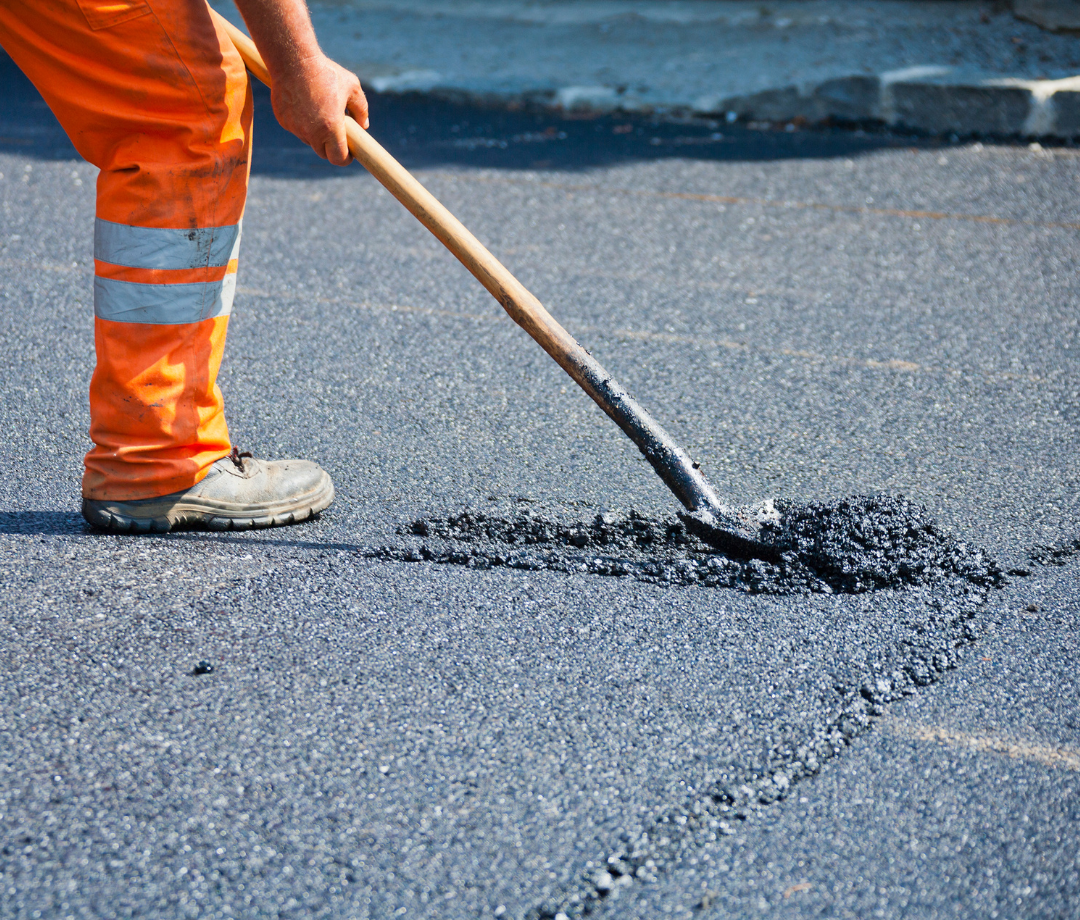 Construction worker using a shovel to spread and level hot asphalt during a road repair project.