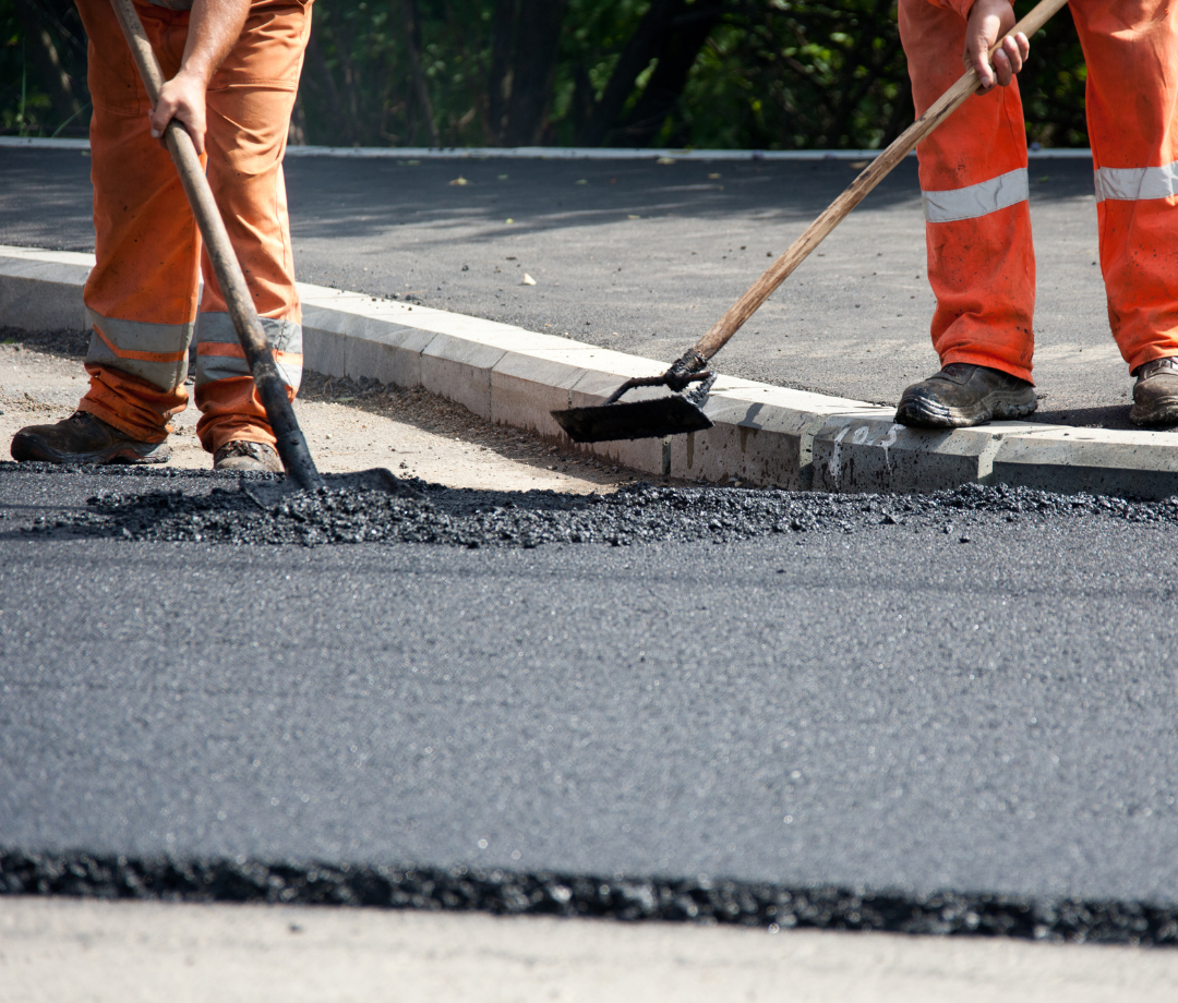 Construction workers using rakes to spread and level hot asphalt along a curb during a road repair project.