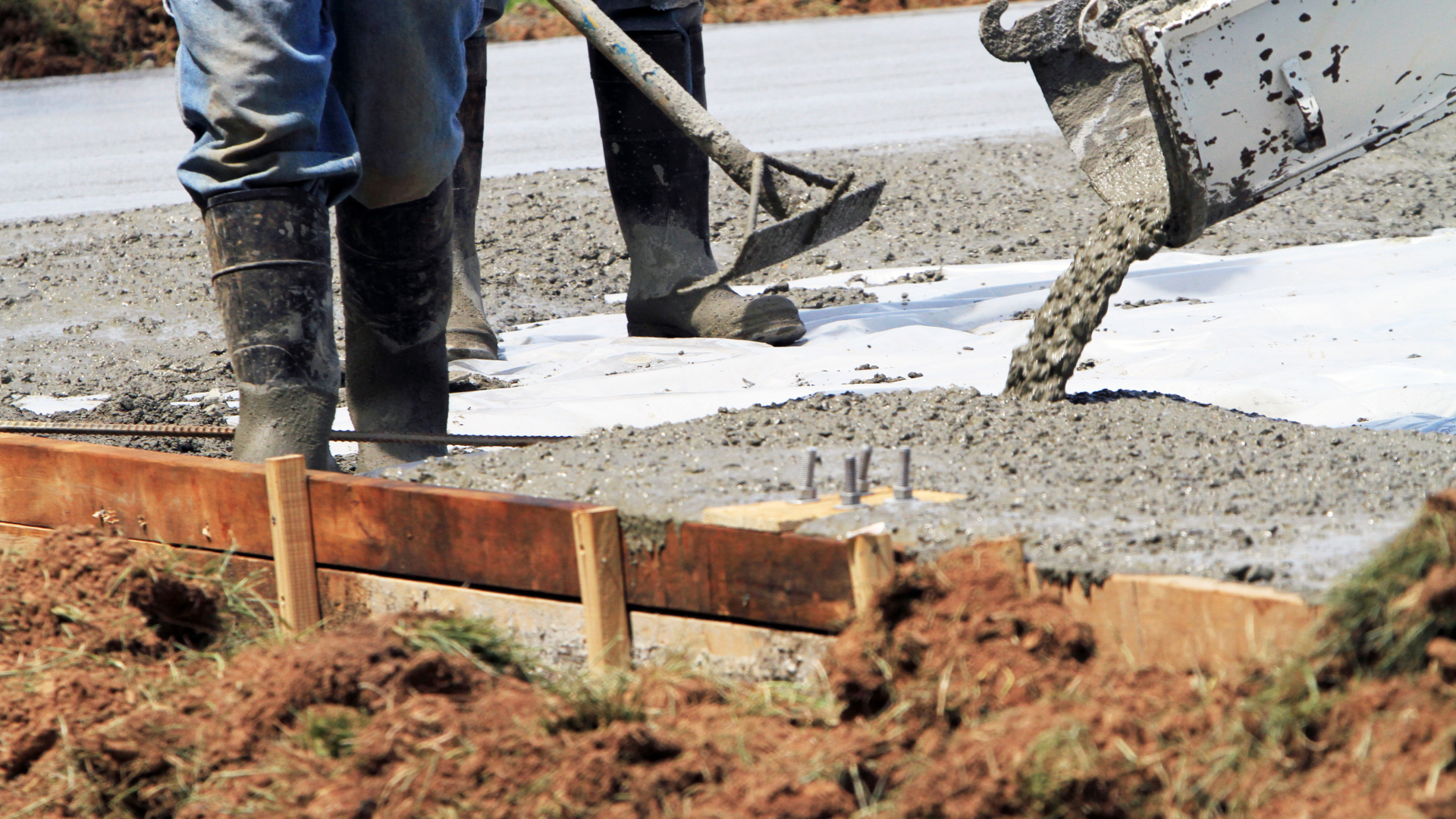 Workers pouring and spreading fresh concrete into wooden forms for a new concrete slab at a construction site.