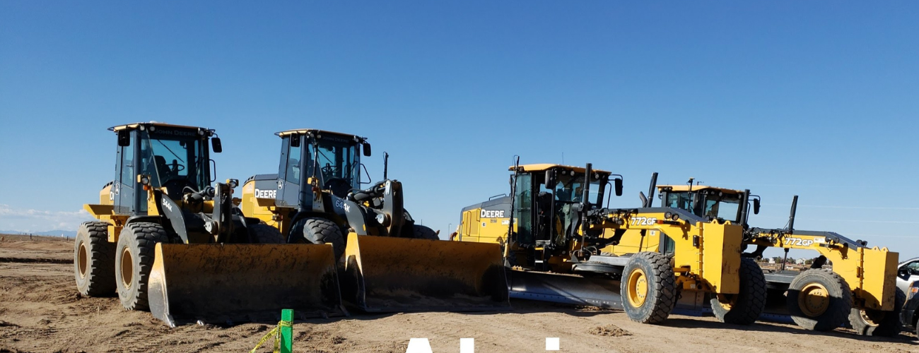 Row of yellow John Deere construction machines, including loaders and motor graders, parked on a dirt construction site under a clear blue sky.