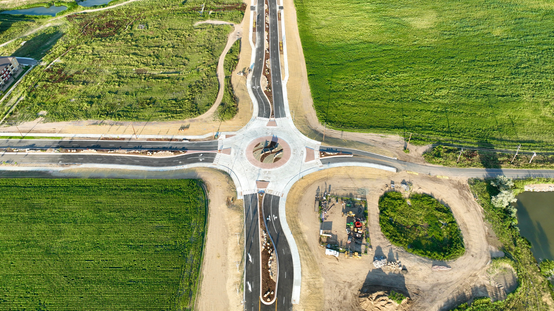 Aerial view of a newly constructed roundabout intersection connecting four paved roads, surrounded by green agricultural fields and open land, with landscaping in the center and construction equipment nearby.