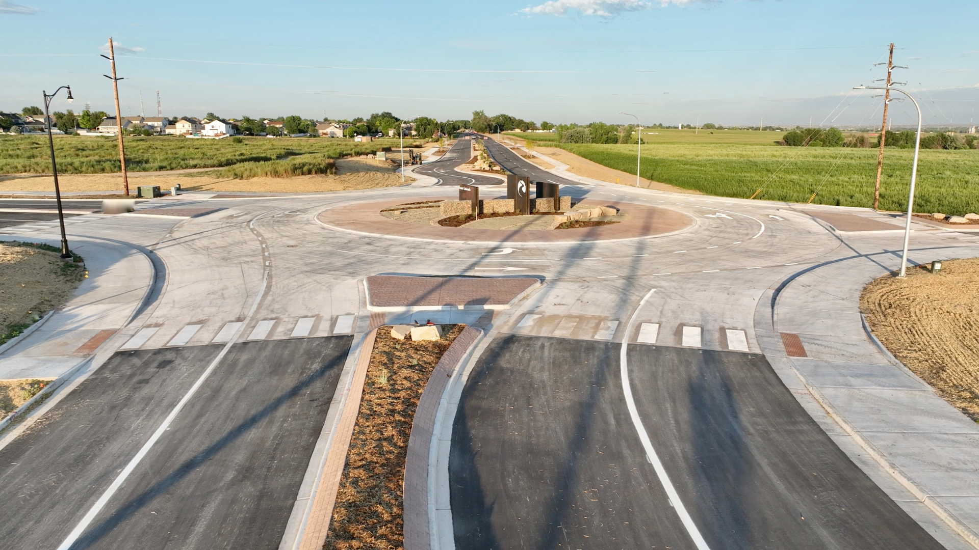 Ground-level view of a newly completed roundabout intersection with marked crosswalks, landscaped center island, and multiple connecting roads, surrounded by open fields and nearby homes.