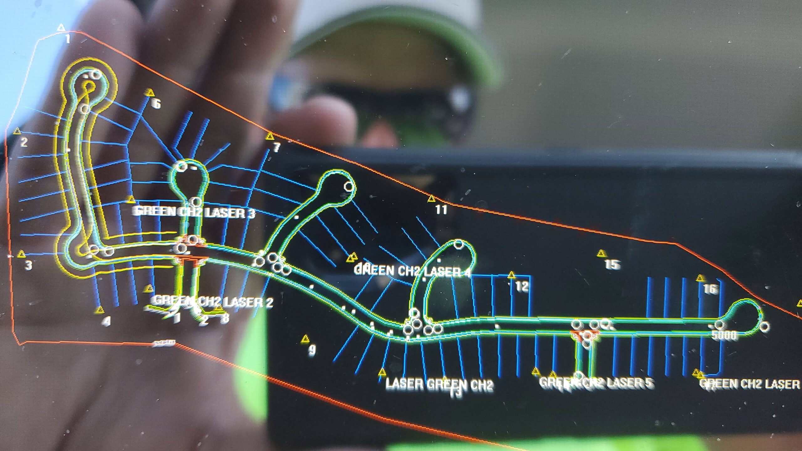 Close-up of a smartphone displaying a digital construction site layout and surveying map, with a worker wearing safety gear visible blurred in the background.