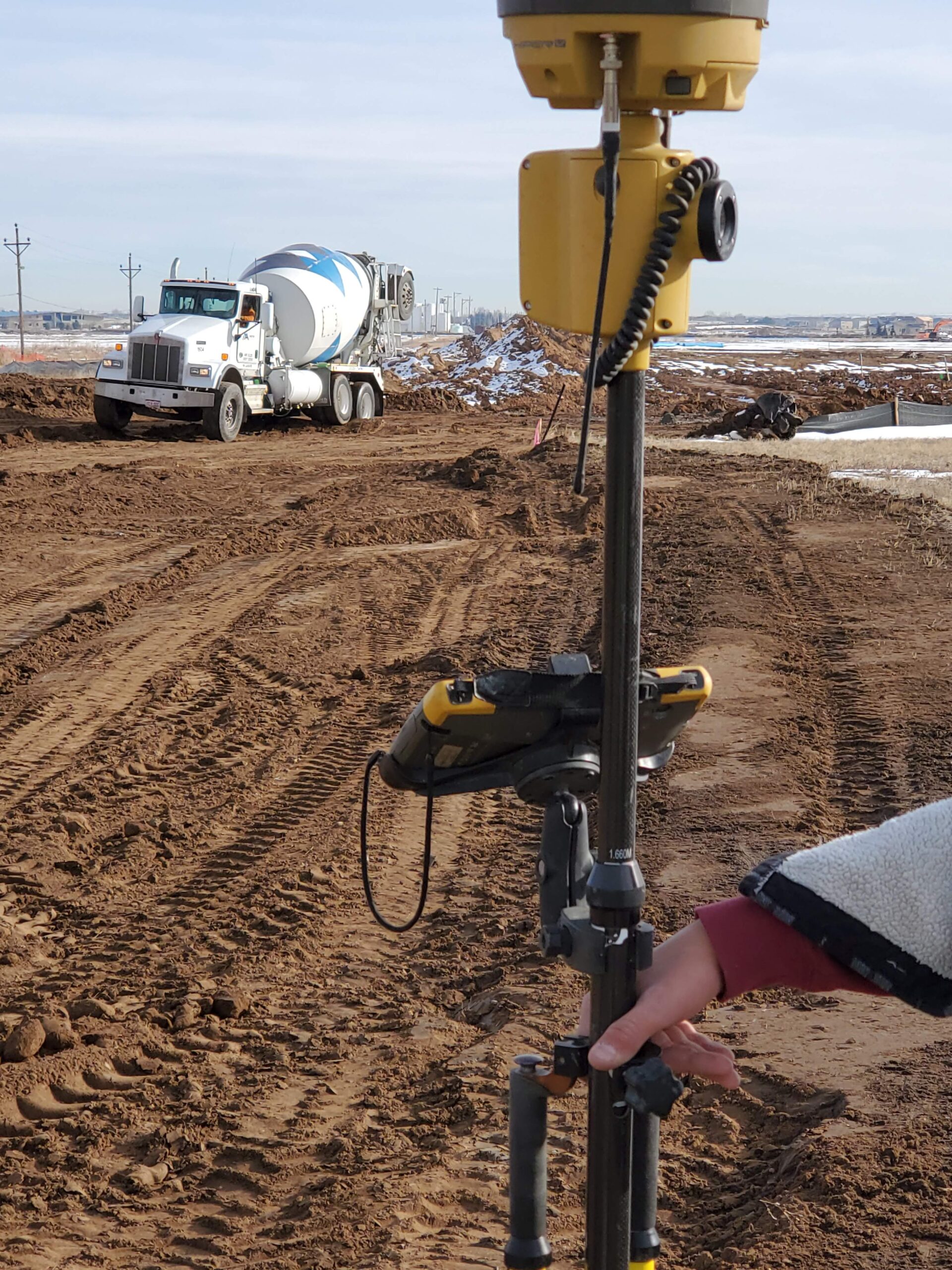 Surveying equipment mounted on a tripod at a construction site while a cement mixer truck drives along a dirt road in the background.