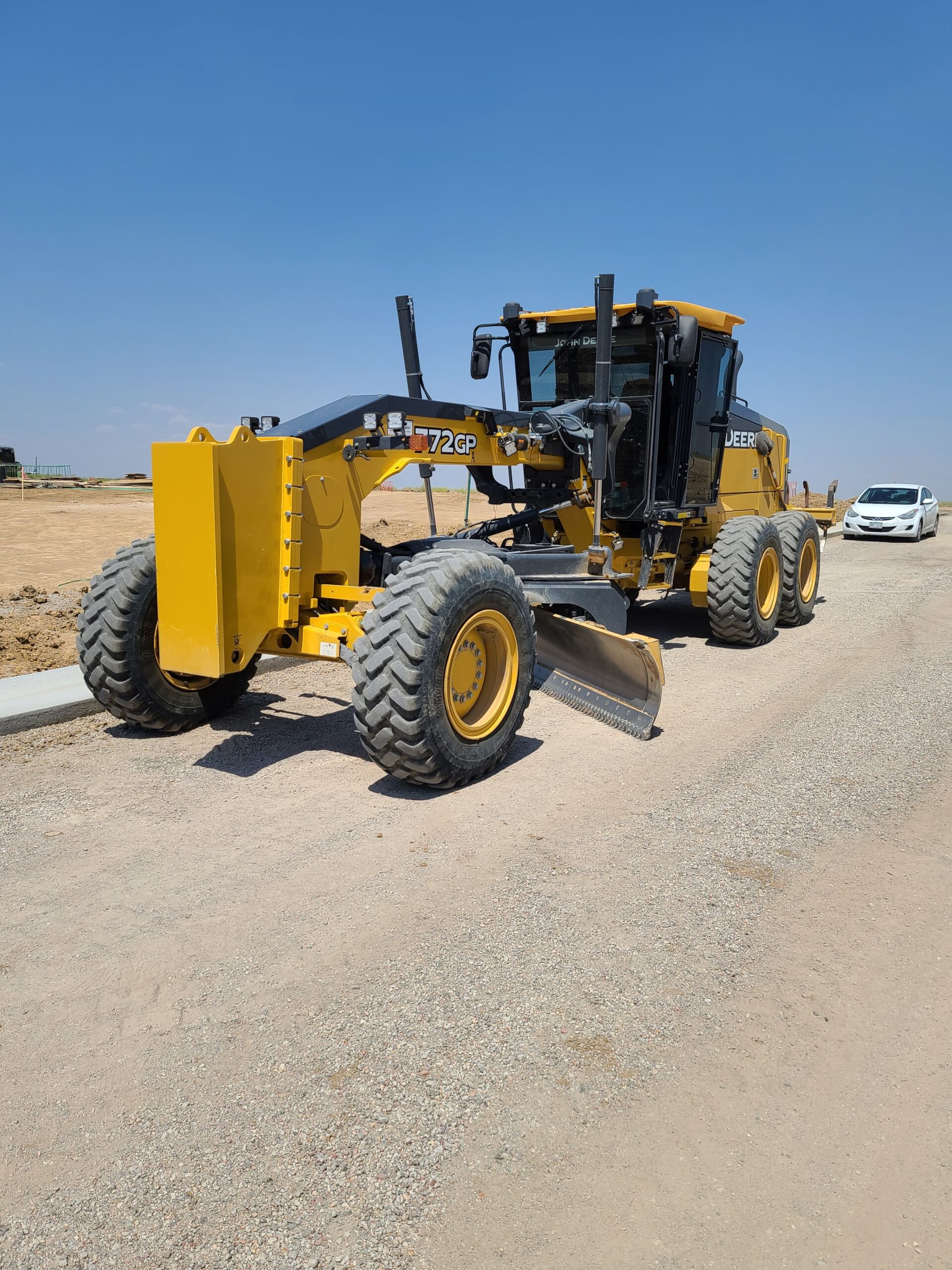 Yellow John Deere 772GP motor grader parked on a gravel road at an active construction site under a clear blue sky.