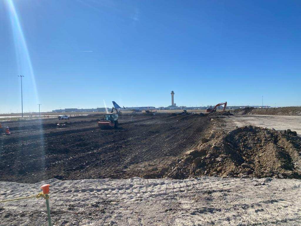 Excavators performing large-scale earthwork near an airport with airplanes and a control tower visible in the background.
