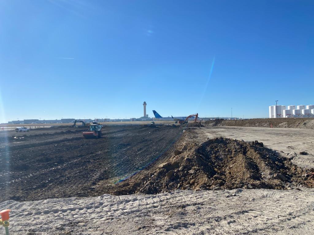 Earthmoving equipment performing large-scale grading at an airport construction site with airplanes and an air traffic control tower visible in the distance.