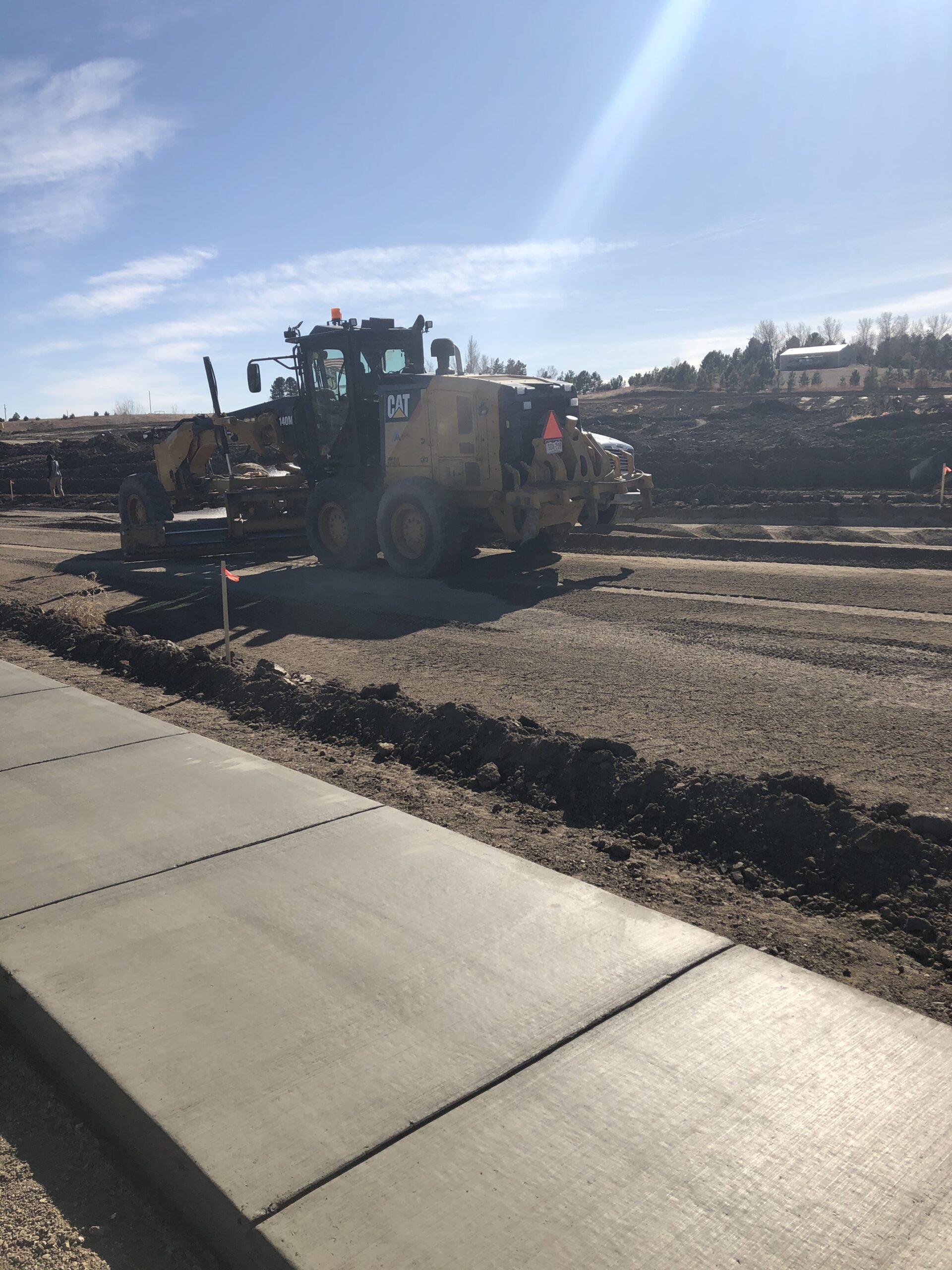Caterpillar motor grader shaping and leveling a dirt roadway beside a newly poured concrete sidewalk at a construction site.