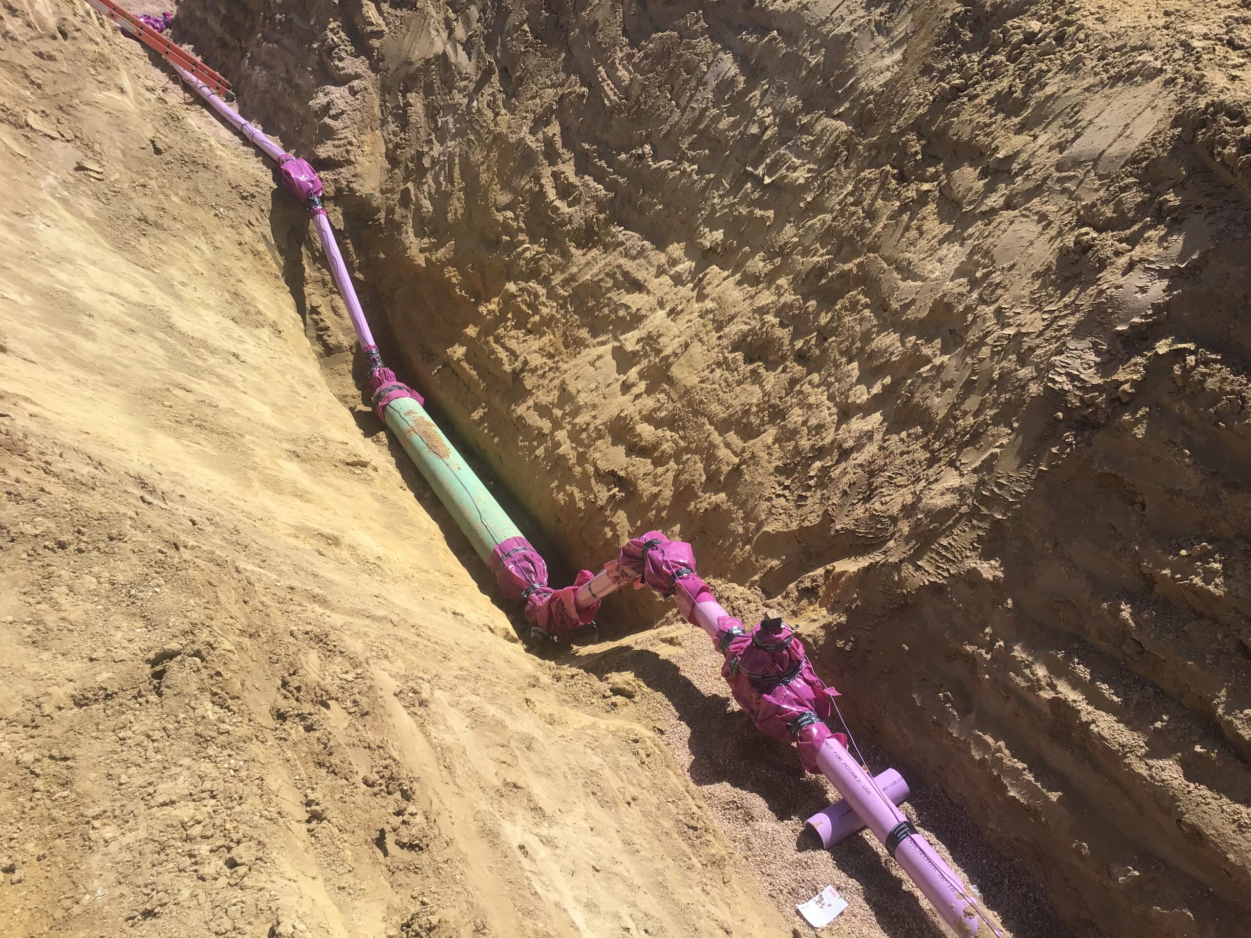 Water utility pipeline installed in a deep excavation trench with joints wrapped for protection and gravel bedding along the pipeline route.