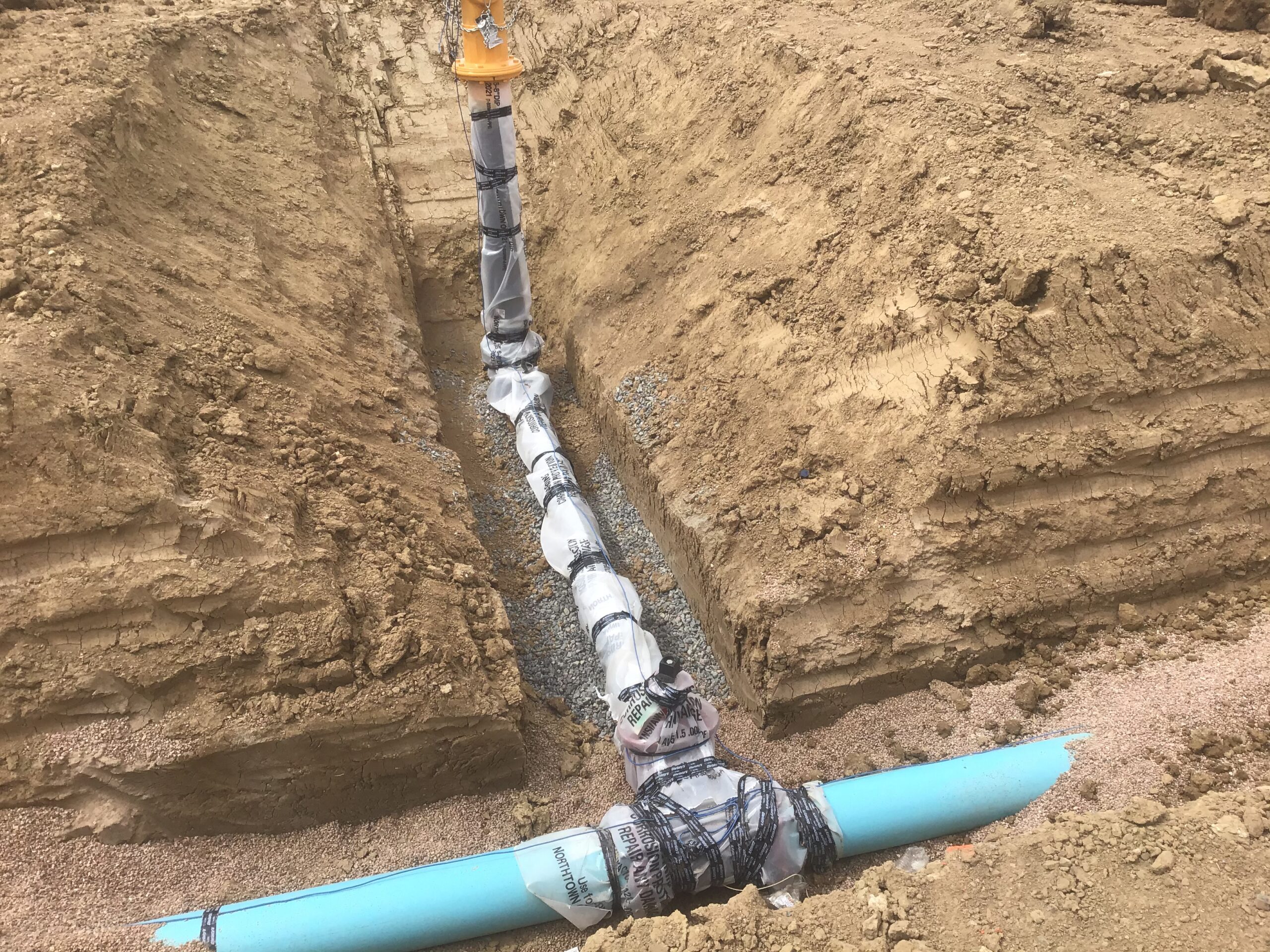 Underground utility installation showing a wrapped vertical pipe connected to a blue water line in a freshly excavated trench with gravel bedding.