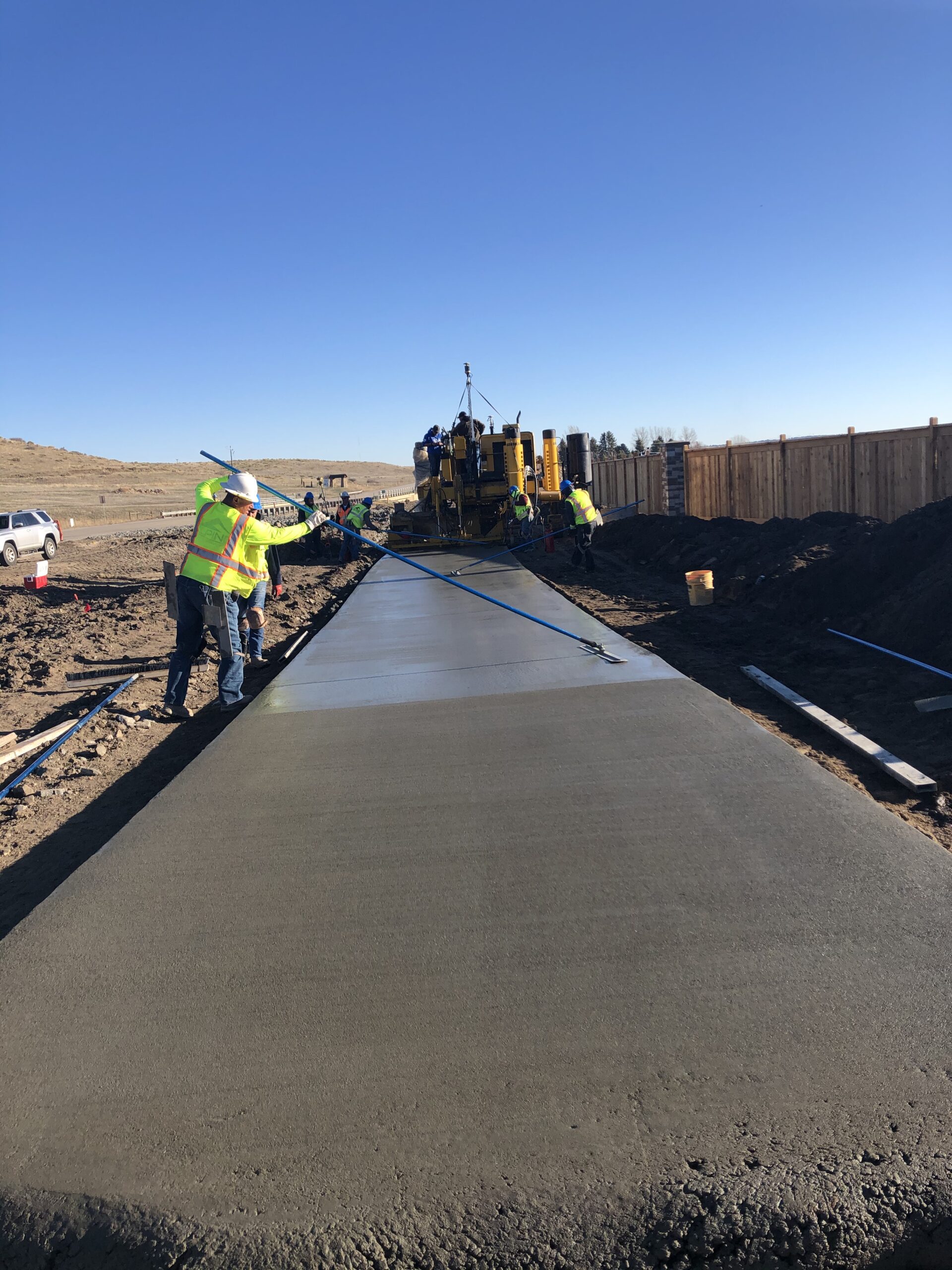 Construction crew finishing a freshly poured concrete roadway using long screeds while a paving machine operates behind them.