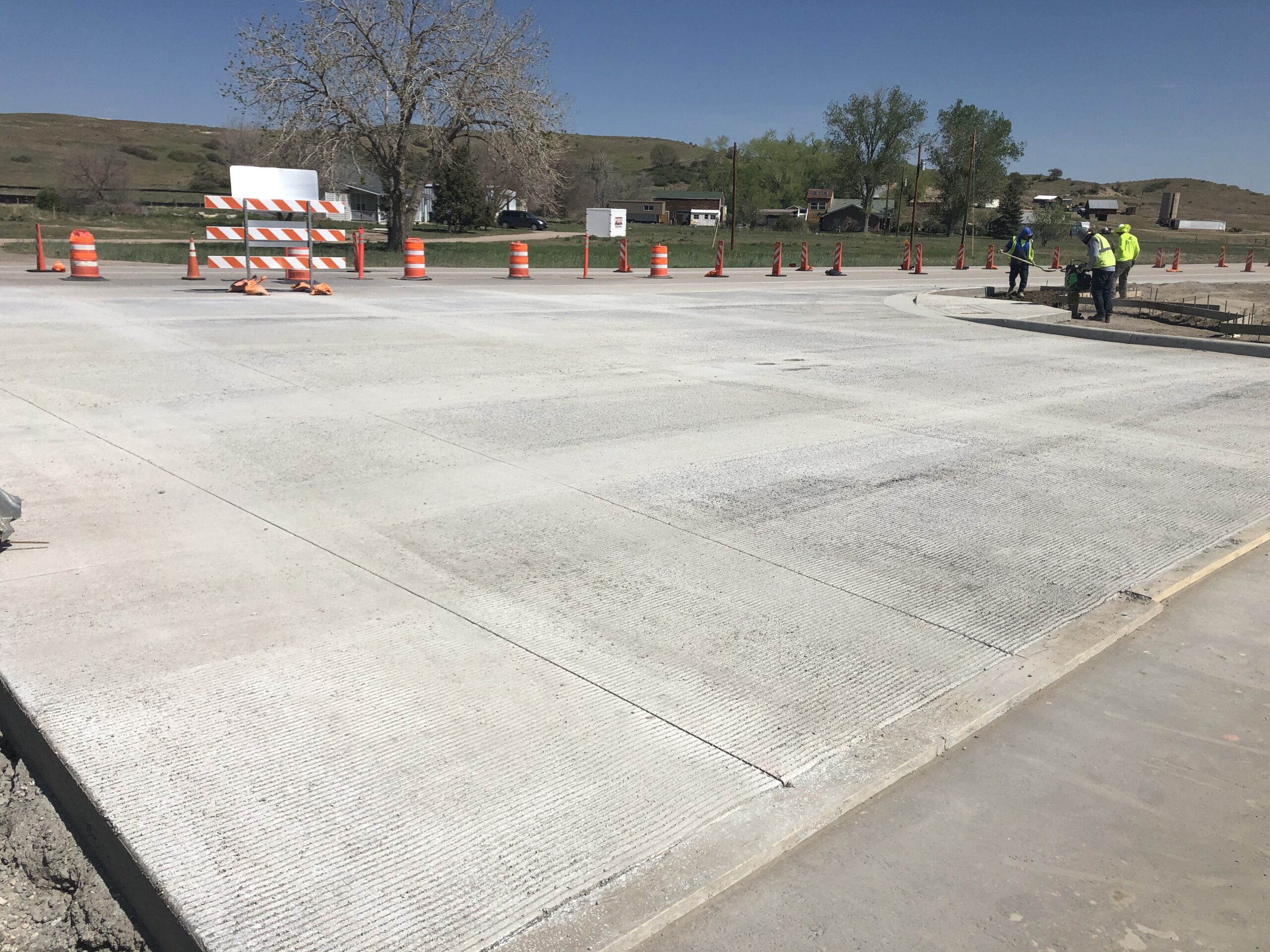 Completed concrete roadway intersection with traffic control barrels and workers performing final construction tasks.