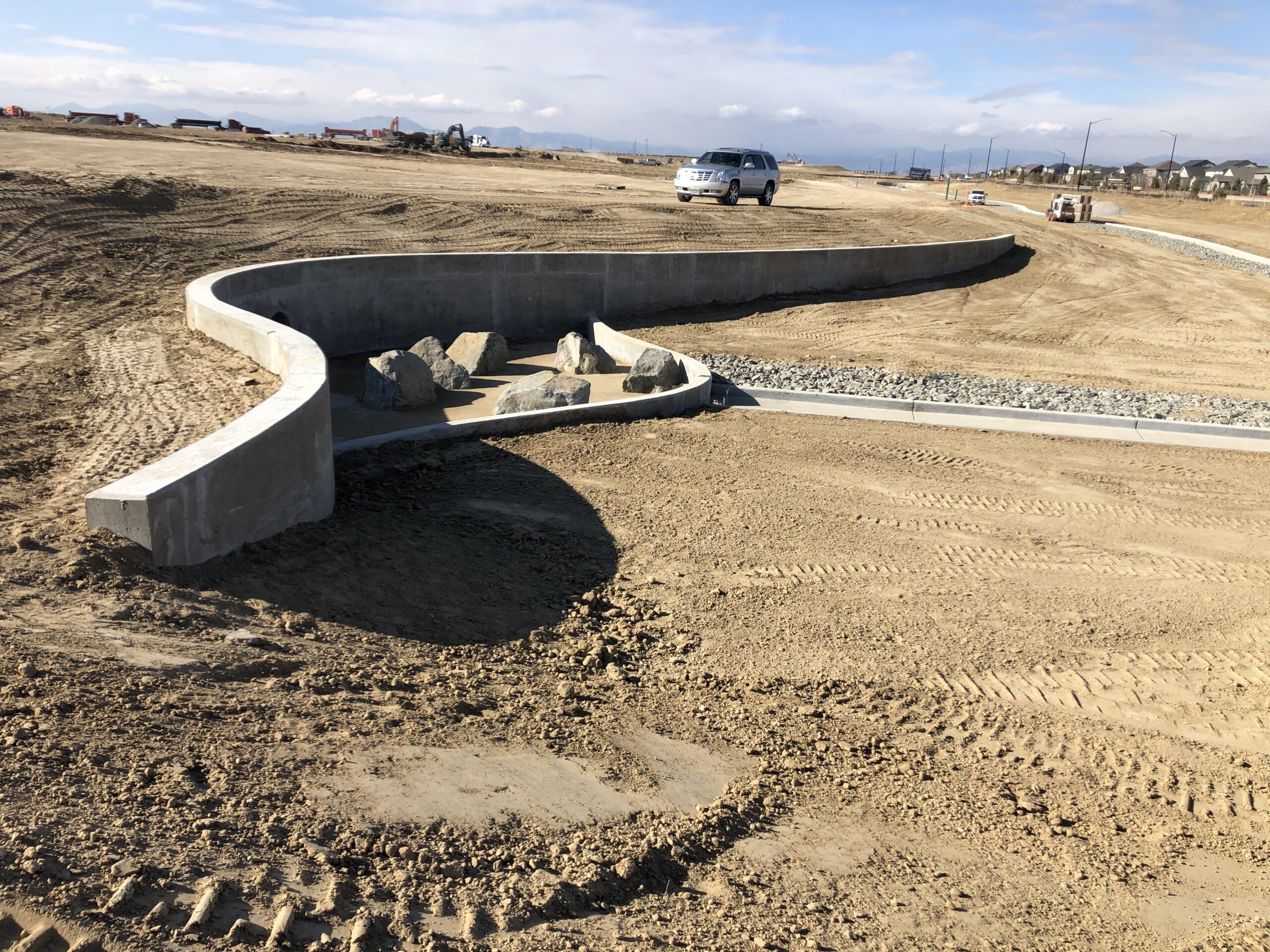 Curved concrete retaining wall and rock-lined drainage feature under construction in a large graded development site with mountains in the distance.