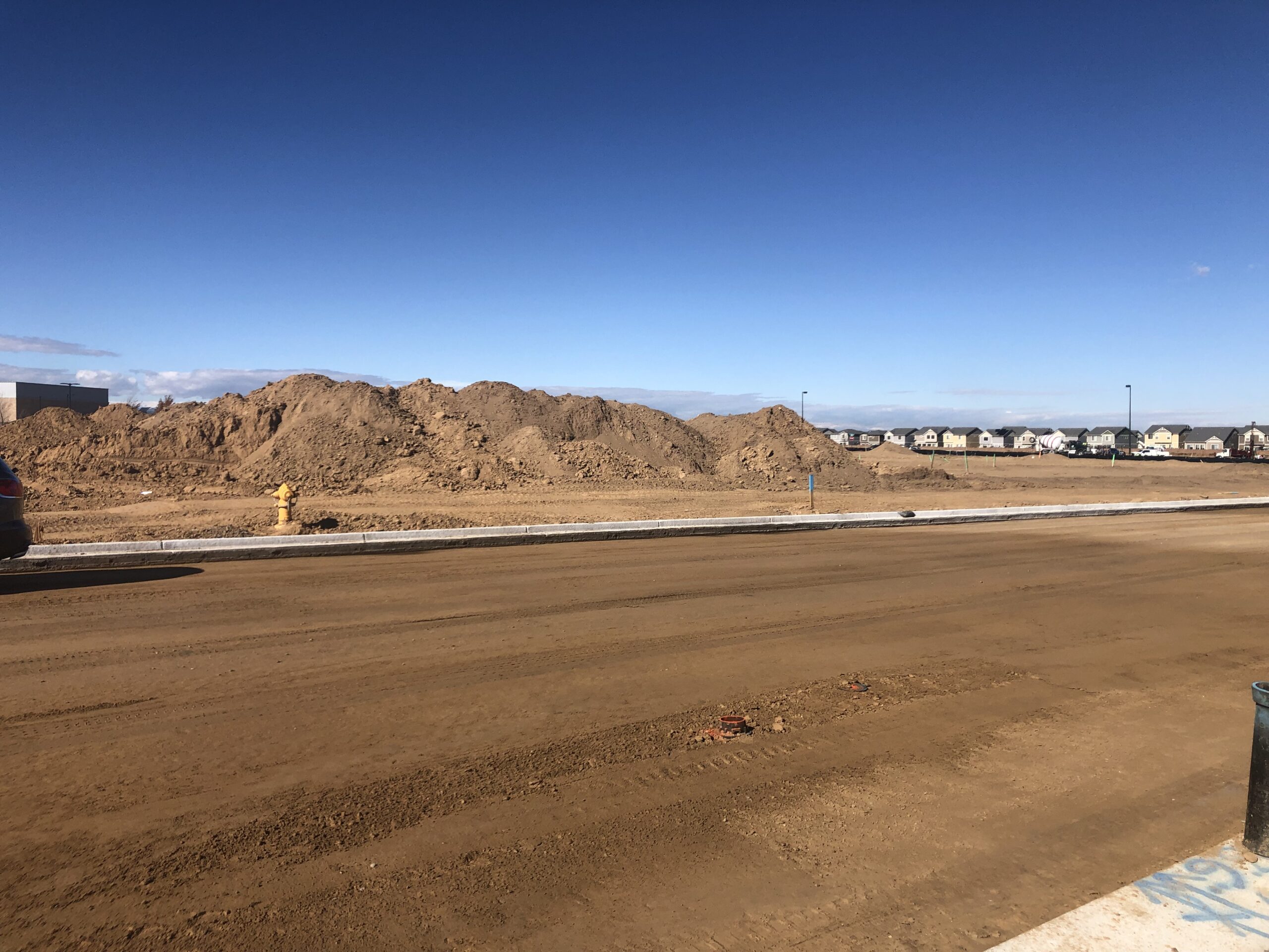 Graded dirt roadway under construction with curb and gutter installed and large soil stockpiles in a developing residential area.