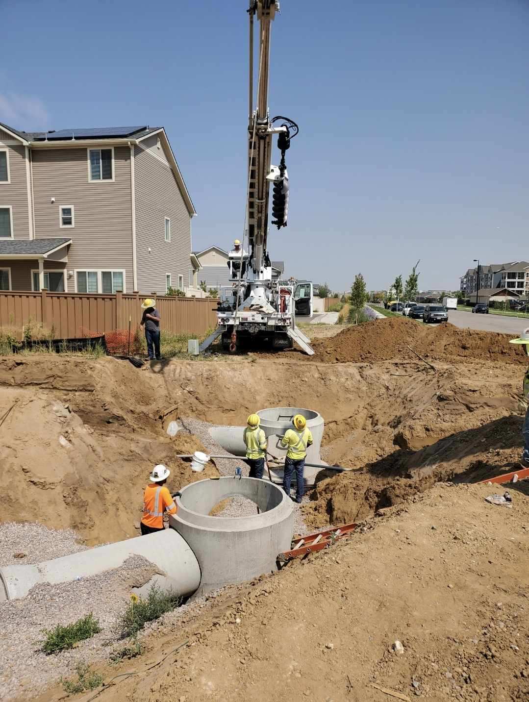 Construction crew installing large precast concrete storm drainage structures in a deep excavation while a crane truck operates beside a residential neighborhood.