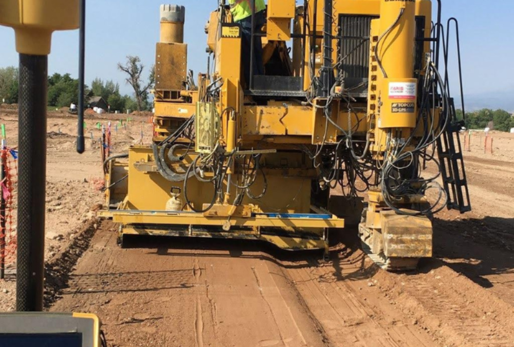 Rear view of a curb and gutter machine shaping concrete along a prepared roadway path at a construction site.
