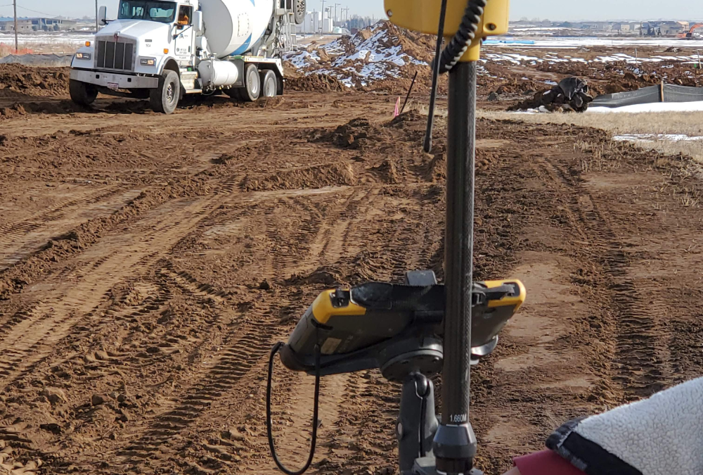 Surveying equipment set up beside a dirt construction roadway with a concrete mixer truck driving through an active site in the background.