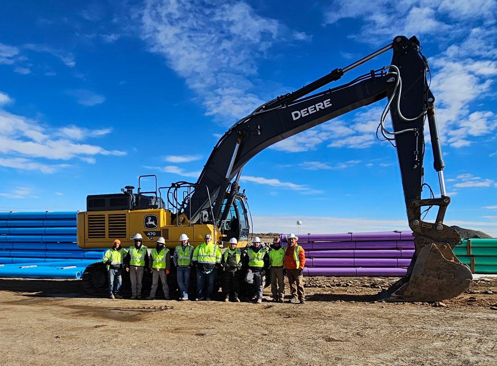 Construction crew standing in front of large excavator and stacked pipes