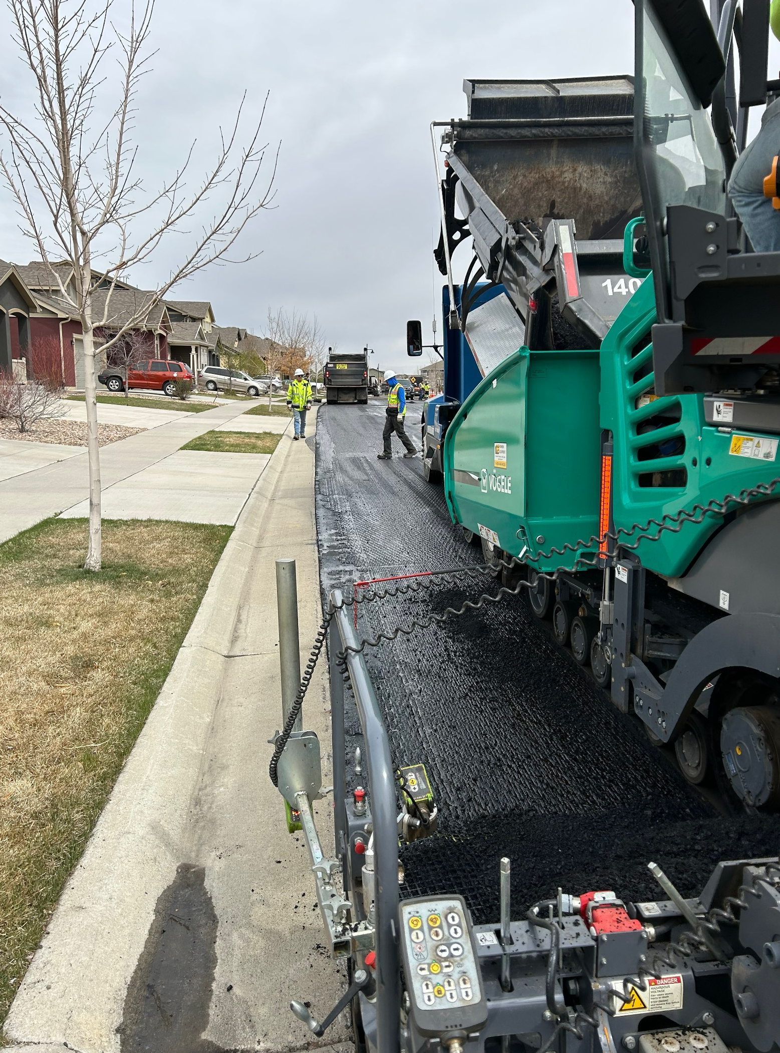 Asphalt paving machine laying fresh asphalt along a residential street while construction workers guide the paving process.