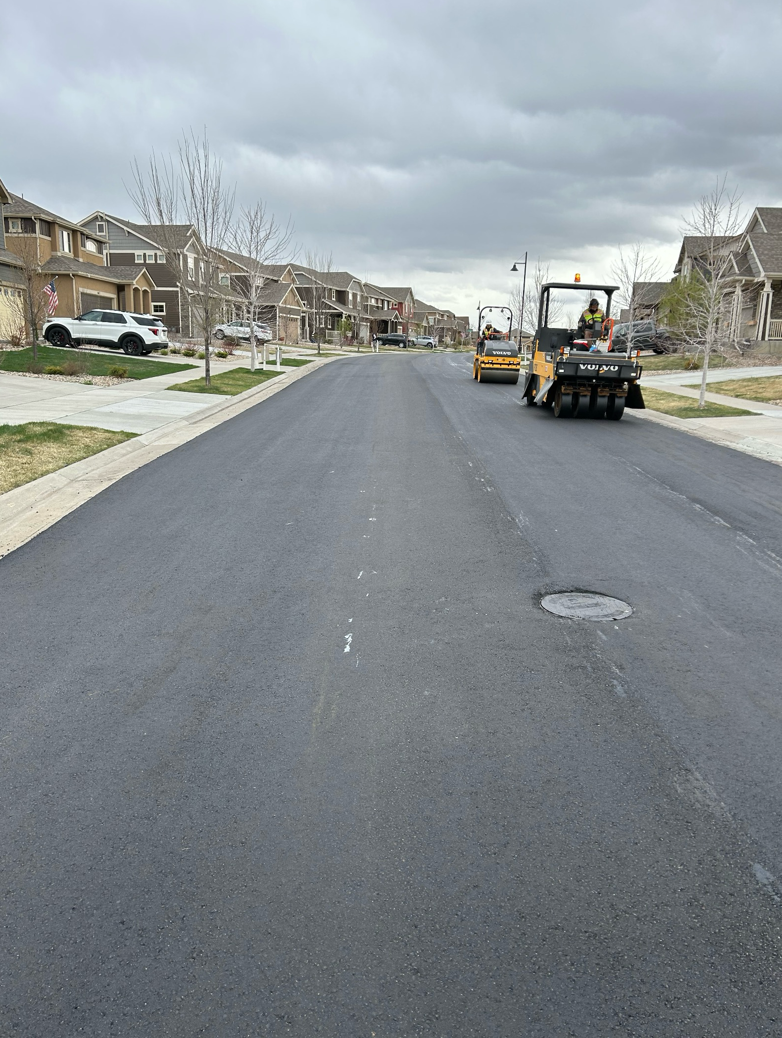 Road rollers compacting freshly paved asphalt on a residential street lined with homes and sidewalks.
