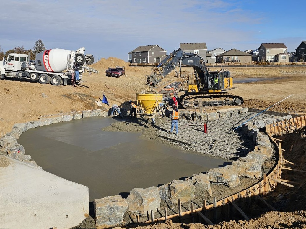 Construction crew pouring and finishing a large circular concrete foundation with an excavator and cement truck nearby.