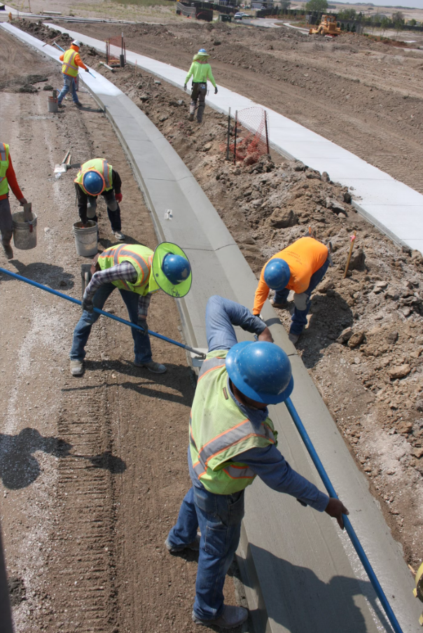 Construction workers wearing hard hats and safety vests install and finish a newly poured concrete curb and sidewalk along a roadside construction project.