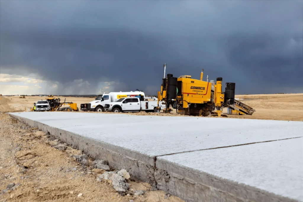 A nearly ground-level shot of a stretch of freshly laid concrete sidewalk on a wide open dirt construction site, with several white trucks and a yellow slipform concrete paver in the background and a dramatic, stormy sky in the distance.