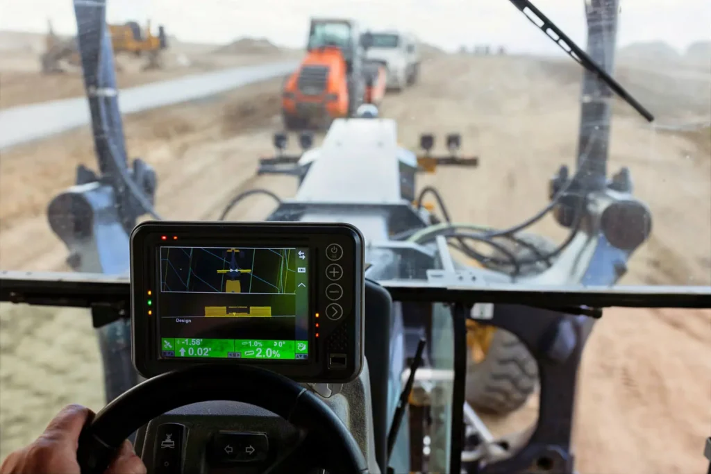 A Topcon GPS machine control display sits above the steering wheel of a piece of heavy machinery being driven across a construction site; other vehicles, a new sidewalk, and an expanse of dirt can be seen through the windshield.