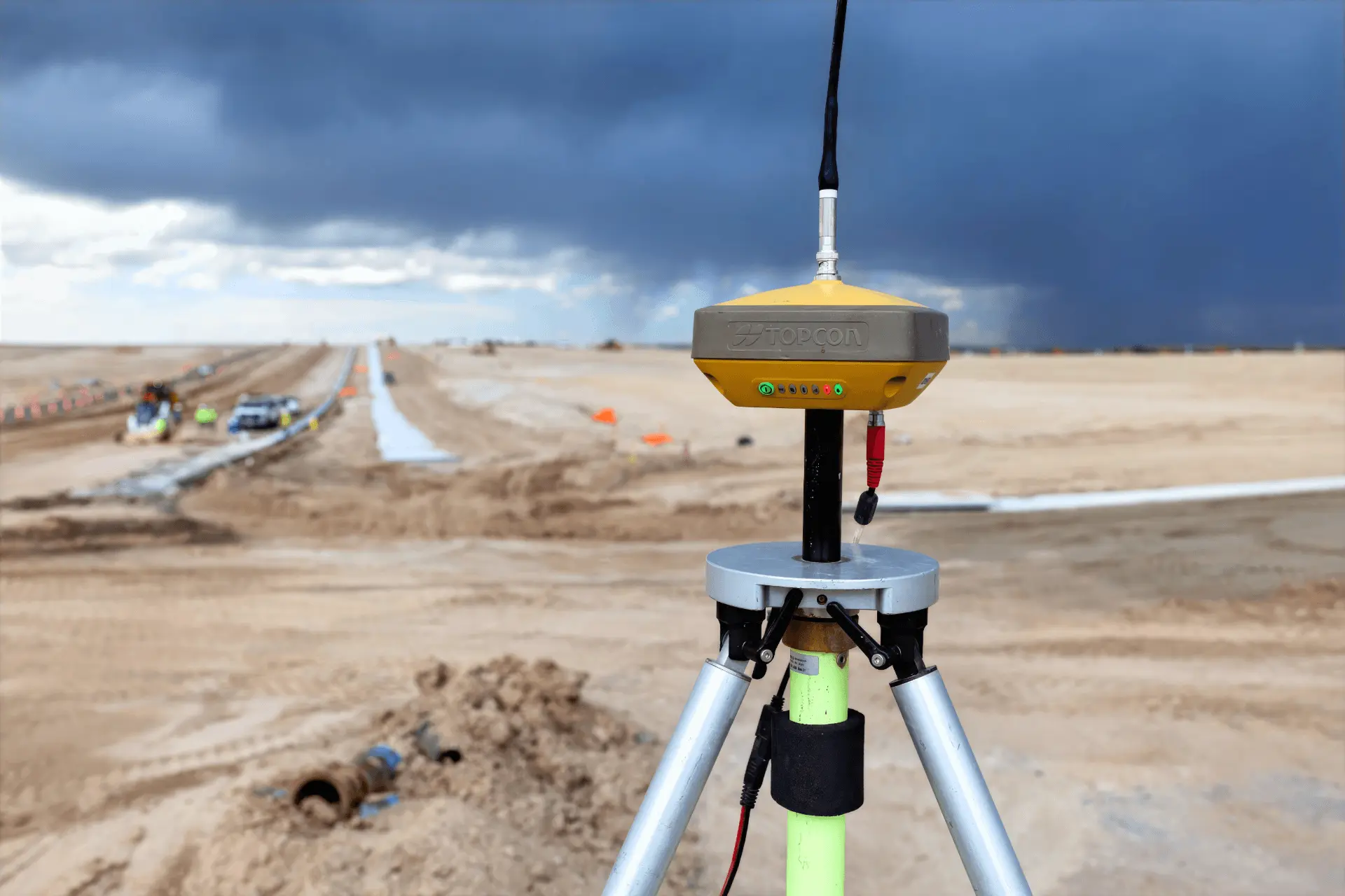 A Topcon GNSS survey rover on a tripod overlooking an Alpine Civil Construction earthwork site, with fresh concrete curb and a stormy Front Range sky
